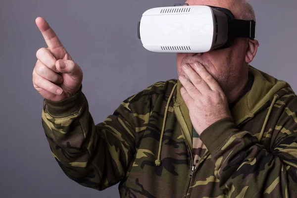 Senior man in wearable technology VR glasses. Confident old man wearing camouflage clothing in virtual reality headset with Interface selective focus hands pointing in the air