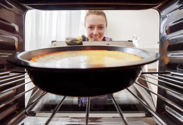 Молода усміхнена домогосподарка дивиться на чізкейк в духовку — стокове фото