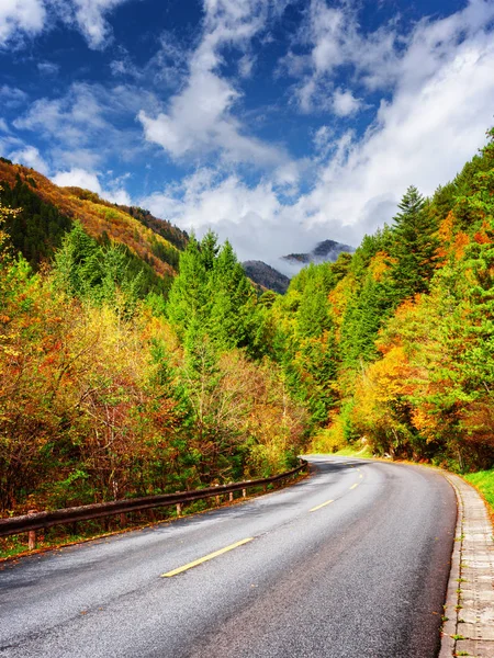 Κάμψη δρόμο μεταξύ πολύχρωμο φθινόπωρο δάσος. Βουνά στο φόντο — Φωτογραφία Αρχείου