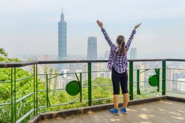 Kollarını havaya kaldırmış genç, güzel bayan turist dağın tepesinden Taipei 'yi seyrediyor. Taipei silueti, Tayvan. Harika bir şehir manzarası. Gökdelenler ve şehir merkezindeki diğer modern binalar.