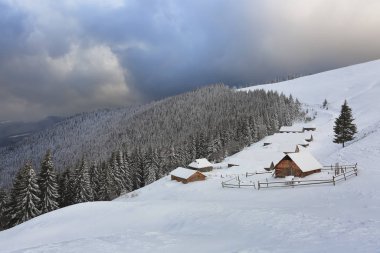 Görkemli kış manzarası. Dramatik bir gökyüzü. Bahçede karla kaplı eski ahşap kulübeler. Yüksek dağların ve ormanların manzarası. Duvar kağıdı arka planı. Konum: Carpathian, Ukrayna, Avrupa.