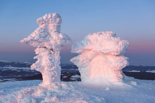 Sněhem pokrytý dům je pokryt ledem, jako marshmallow, ozářený jemným růžovým světlem zapadajícího slunce. Horská krajina v pozadí. — Stock fotografie