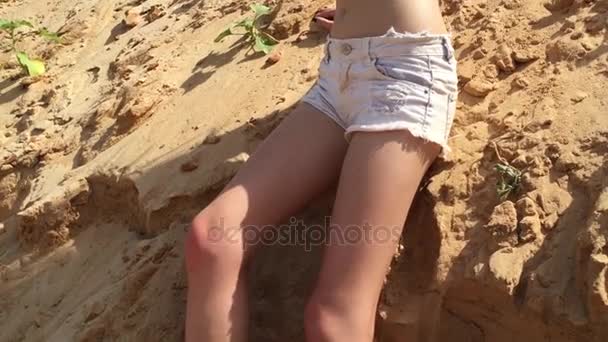 Modelo de mujer de moda con el pelo largo en pantalones cortos de mezclilla y una camiseta, posando sexy sobre el telón de fondo de dunas de arena en el desierto . — Vídeos de Stock