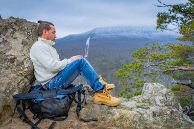 Hipster adam defteriyle çalışıyor. Kayalık dağda oturuyor. Manzaralı bir arka planda.