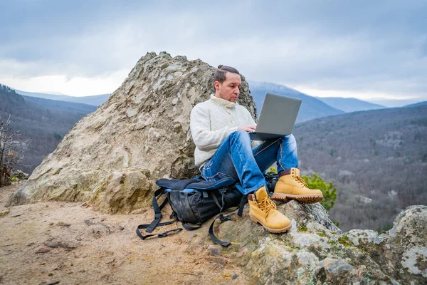 Hipster człowiek pracuje z notebooka siedzi na skalistej górze na pięknym tle malowniczego klifu — Zdjęcie stockowe