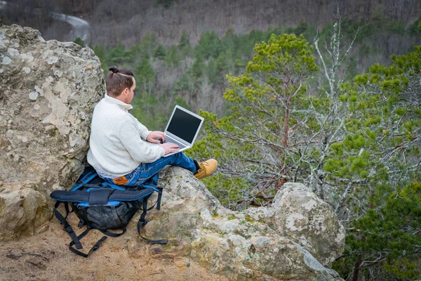 Hombre de viaje que trabaja con el ordenador portátil sentado en la montaña rocosa en el hermoso fondo del acantilado escénico — Foto de Stock