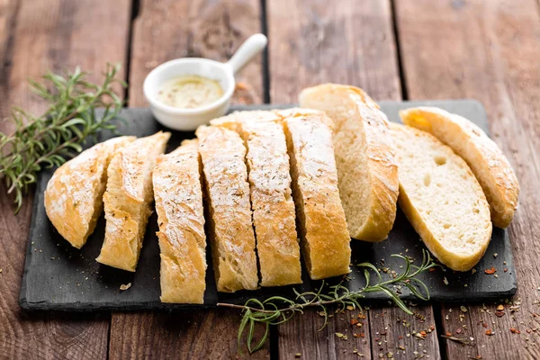 Pão de ciabatta caseiro fresco fatiado em tábua de ardósia de pedra, azeite e alecrim em fundo de madeira — Fotografia de Stock