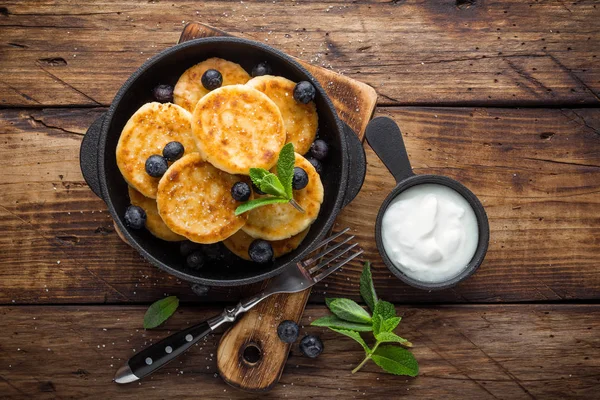 Köstliche Quark-Pfannkuchen oder Syrniki mit frischen Blaubeeren in gusseiserner Pfanne auf dunklem rustikalem Holzhintergrund, über der Ansicht. Leckeres Frühstück. — Stockfoto