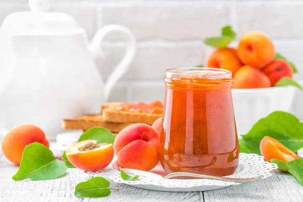 Apricot jam in a jar and fresh fruits with leaves on white wooden table, breakfast — Stock Photo, Image