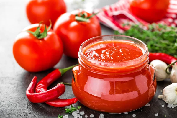 Tomato paste, puree in glass jar and fresh tomatos on dark background. Hot vegetable sauce with chili pepper and tomatoes