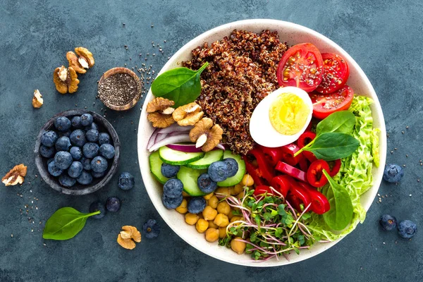 Buddha bowl dinner with boiled egg, chickpea, fresh tomato, sweet pepper, cucumber, savoy cabbage, red onion, green sprouts, spinach leaves, blueberry, walnuts, chia and quinoa. Healthy dish, lunch bowl. Detox diet. Balanced food. Top view