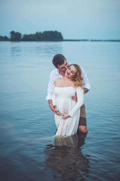 年轻英俊的男人和他怀孕的妻子站在湖边的水和拥抱 — 图库照片