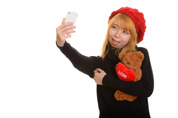 年轻快乐的亚洲女人笑着抱着泰迪熊 一边带着爱心和爱心 一边用手机自拍 — 图库照片