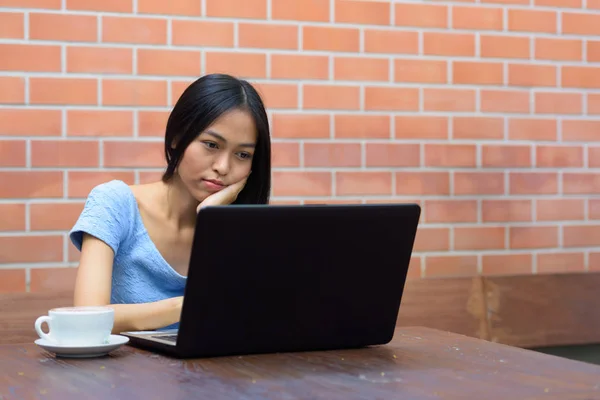 Jeune belle adolescente asiatique reposant menton sur la main tout en utilisant un ordinateur portable avec cappuccino sur une table en bois contre un mur de briques — Photo