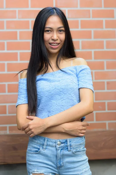 Jovem feliz menina adolescente asiática sorrindo com os braços cruzados contra a parede de tijolo — Fotografia de Stock