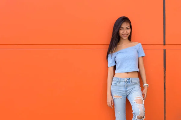 Jeune fille asiatique heureuse adolescente souriant tout en s'appuyant contre le mur peint orange — Photo