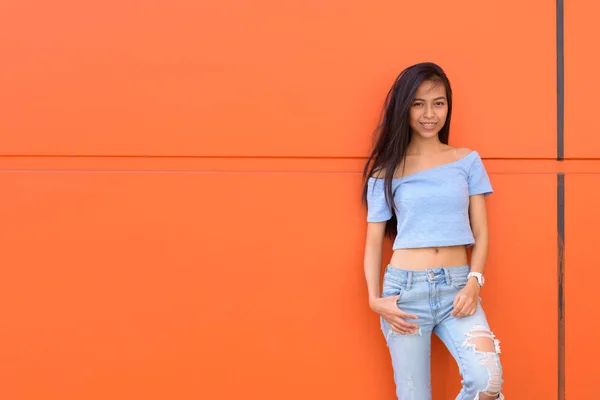 Joven feliz asiática adolescente sonriendo mientras posa y se apoya contra la pared pintada de naranja —  Fotos de Stock