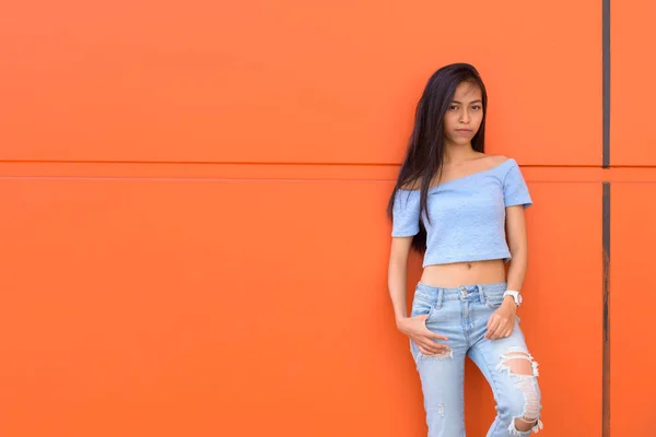 Fiatal gyönyörű ázsiai tizenéves lány jelentő és támaszkodva narancssárga festett fal — Stock Fotó