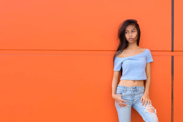 Fiatal gyönyörű ázsiai tizenéves lány gondolkodás és a távolság, míg narancssárga festett fal támaszkodva néztem — Stock Fotó