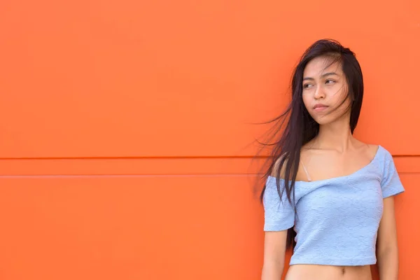 Junge schöne asiatische Teenager-Mädchen denken, während sie sich gegen orange bemalte Wand lehnt — Stockfoto