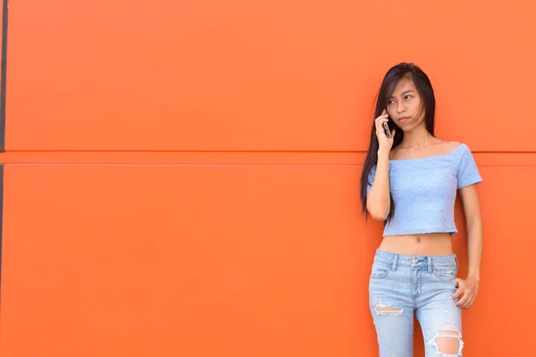 Young beautiful Asian teenage girl talking on mobile phone while standing and thinking against orange painted wall — Stock Photo, Image