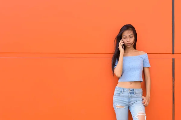 Fiatal gyönyörű ázsiai tizenéves lány beszél mobiltelefonon, miközben gondolkodás és a narancssárga festett falnak lenézett — Stock Fotó