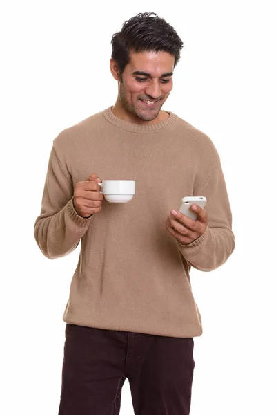 Young happy Persian man using mobile phone while holding coffee — Stock Photo, Image