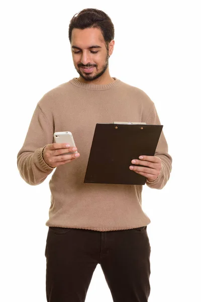 Young happy Indian man holding clipboard while using mobile phon — Stock Photo, Image