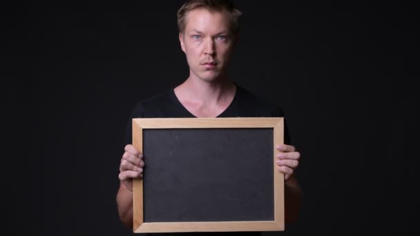 Young handsome man holding blackboard against black background — Stock Video