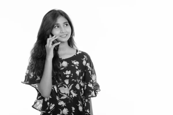 Studio shot of young happy Persian woman smiling and thinking while talking on mobile phone — Stock Photo, Image