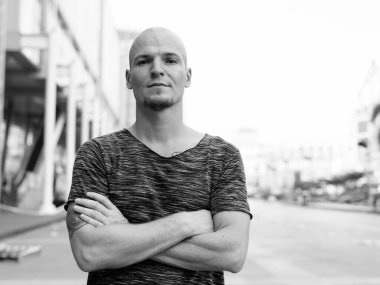 Şehirdeki alışveriş merkezinin dışında kolları çaprazlanmış genç ve kel bir adam.