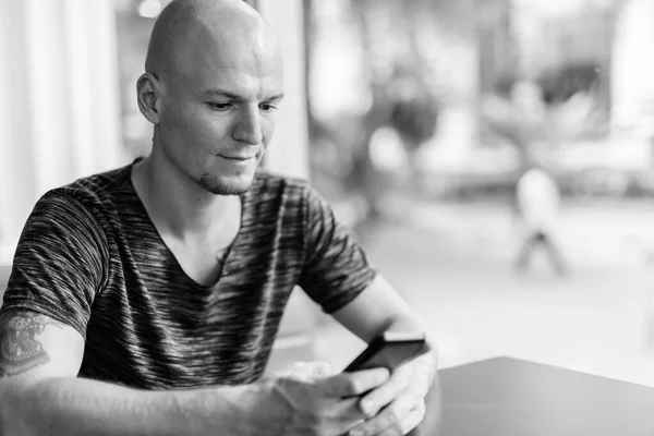 Young handsome bald man using mobile phone inside restaurant with glass windows — Stock Photo, Image