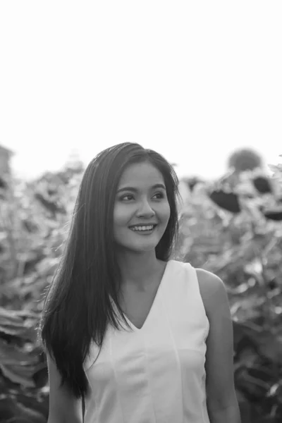 Joven mujer asiática feliz sonriendo y pensando mientras mira a la distancia en el campo de los girasoles florecientes —  Fotos de Stock