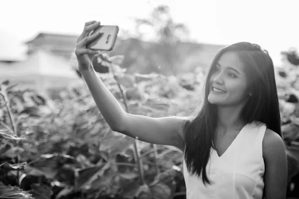 Young happy Asian woman smiling while taking selfie picture with mobile phone in the field of blooming sunflowers — Stock Photo, Image