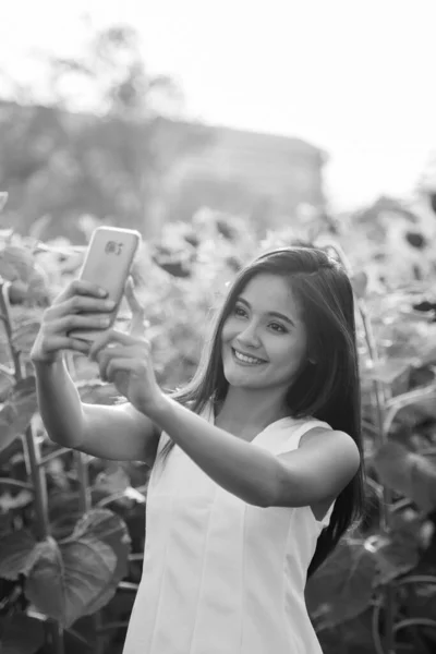 Junge glückliche Asiatin lächelt beim Selfie-Foto mit Handy im Feld blühender Sonnenblumen — Stockfoto