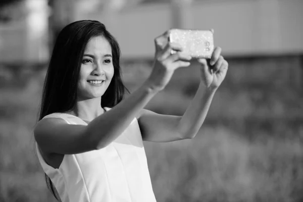 Joven mujer asiática feliz sonriendo mientras toma foto selfie con teléfono móvil contra el campo de hierba —  Fotos de Stock