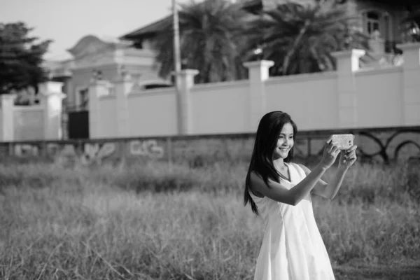 Young happy Asian woman smiling while taking selfie picture with mobile phone against grass field and fancy house — Stock Photo, Image