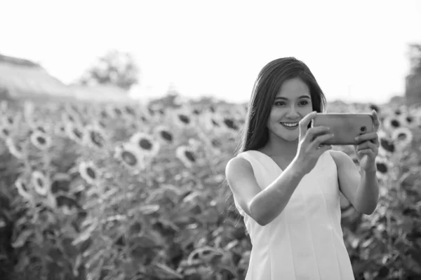 Young happy Asian woman smiling while taking selfie picture with mobile phone in the field of blooming sunflowers — Stock Photo, Image