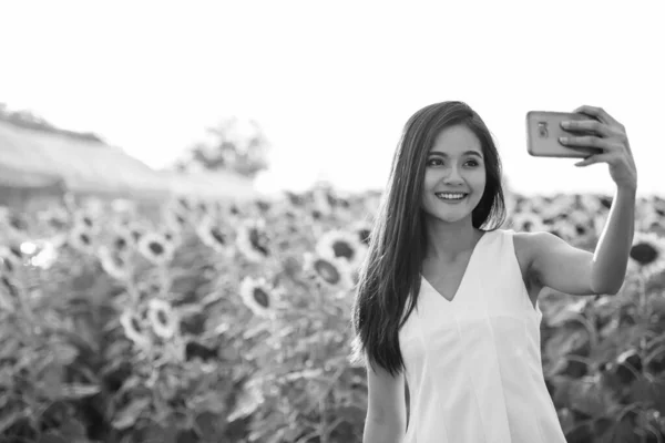 Young happy Asian woman smiling while taking selfie picture with mobile phone in the field of blooming sunflowers — Stock Photo, Image