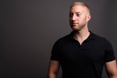 Stüdyo resmi, sarı saçlı, siyah polo tişörtlü, gri arka planda bir adam.
