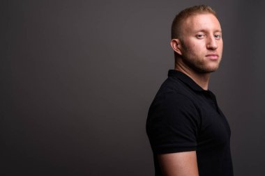 Stüdyo resmi, sarı saçlı, siyah polo tişörtlü, gri arka planda bir adam.