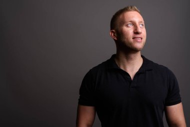 Stüdyo resmi, sarı saçlı, siyah polo tişörtlü, gri arka planda bir adam.