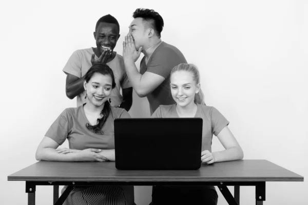 Studio shot of happy diverse group of multi ethnic friends souriant tout en utilisant un ordinateur portable sur une table en bois avec des amis chuchotant à l'arrière — Photo