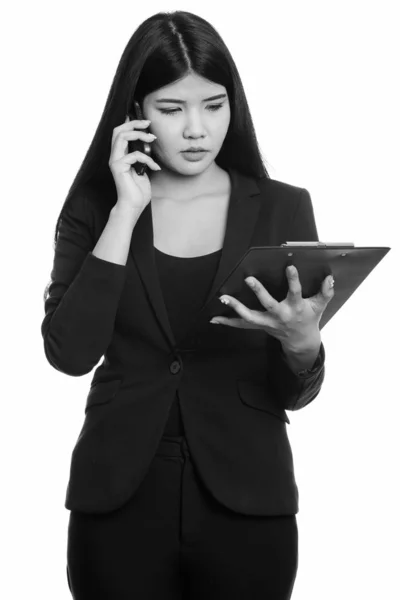 Estúdio tiro de jovem empresária asiática lendo na área de transferência enquanto fala no telefone móvel — Fotografia de Stock