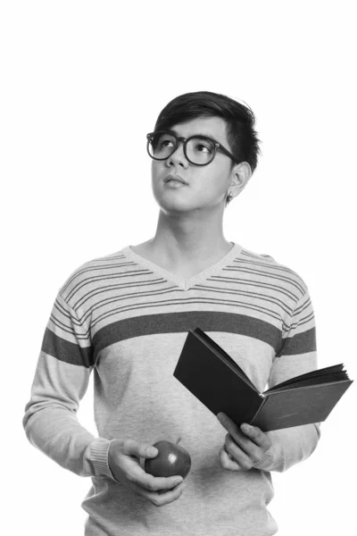 Studio shot van jong knap aziatisch man holding boek en rode appel terwijl denken — Stockfoto