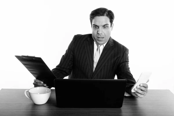 Empresário persa chocado segurando telefone celular e área de transferência ao usar laptop com xícara de café na mesa de madeira — Fotografia de Stock