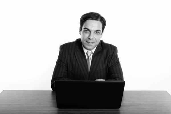 Studioaufnahme eines jungen persischen Geschäftsmannes, der mit Laptop auf einem Holztisch vor weißem Hintergrund sitzt — Stockfoto