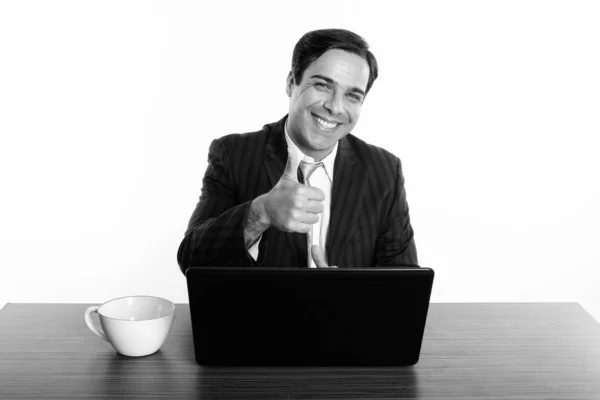 Estudio de toma de feliz hombre de negocios persa sonriendo y dando pulgar hacia arriba con el ordenador portátil y taza de café en la mesa de madera —  Fotos de Stock