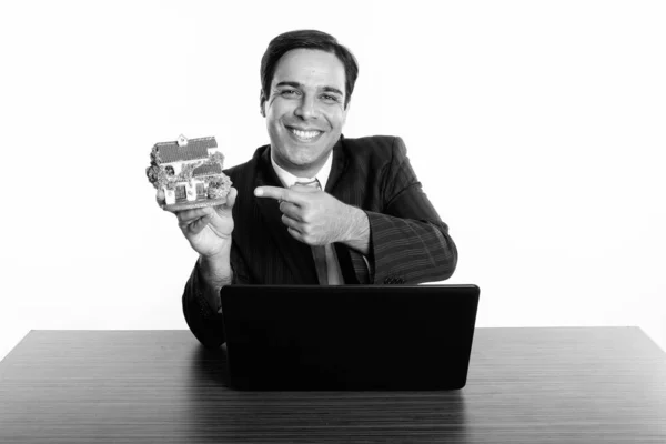 Feliz hombre de negocios persa sosteniendo y señalando la figura de la casa con el ordenador portátil en la mesa de madera —  Fotos de Stock