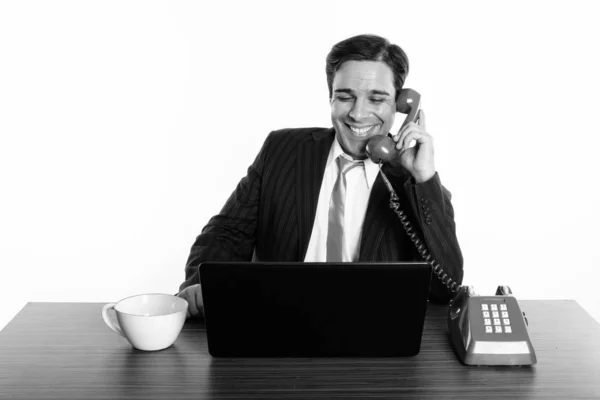 Szczęśliwy perski biznesmen uśmiecha się i rozmawia przez stary telefon podczas korzystania z laptopa z filiżanką kawy na drewnianym stole — Zdjęcie stockowe